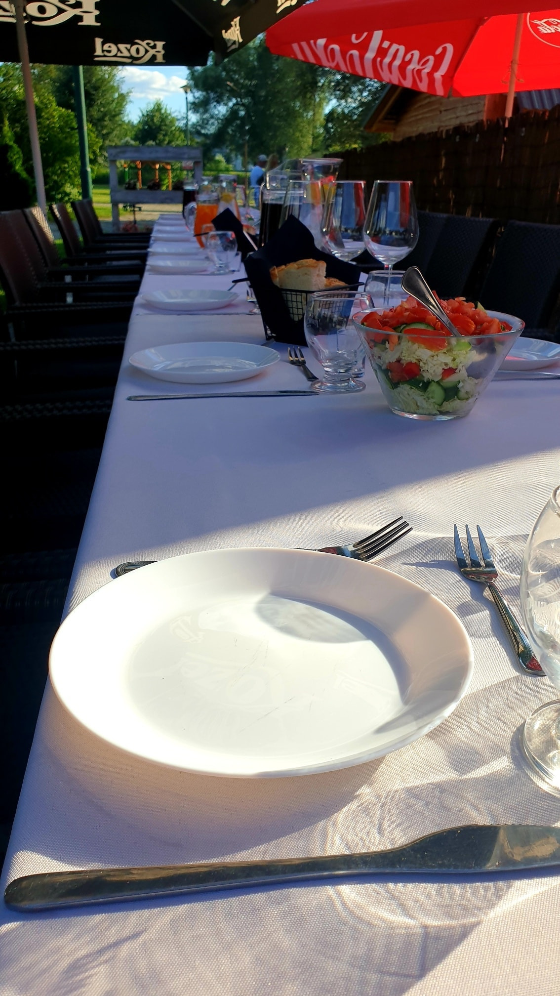 Długi stół nakryty białym obrusem, z talerzami, sztućcami, szklankami, koszyczkiem z pieczywem i miską sałatki, ustawiony na zewnątrz pod parasolami, przygotowany dla gości.