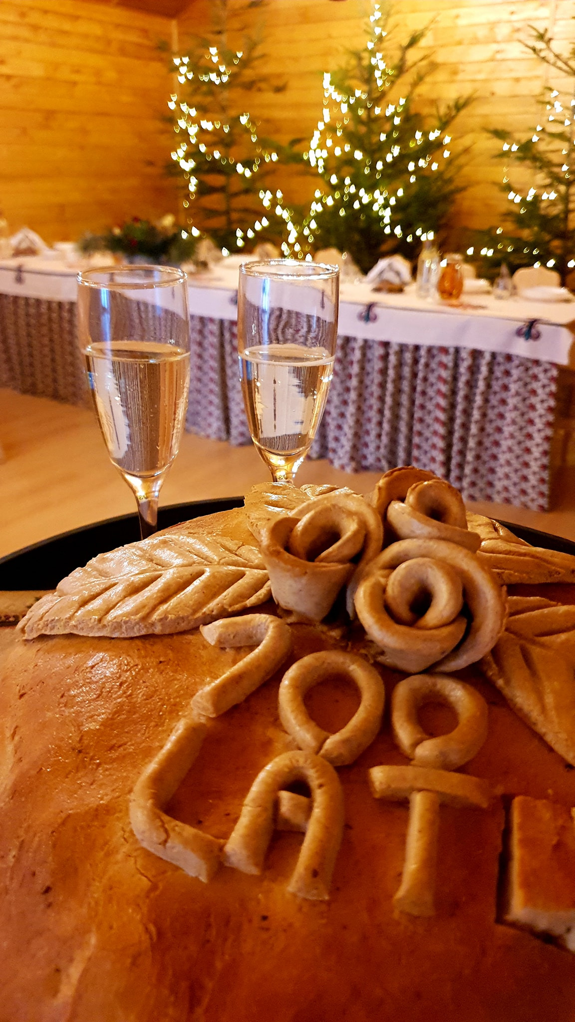 Udekorowany chleb okolicznościowy z napisem '200 LAT!', kwiatami z ciasta i dwoma kieliszkami szampana w tle, świątecznie oświetlone choinki i nakryty stół.