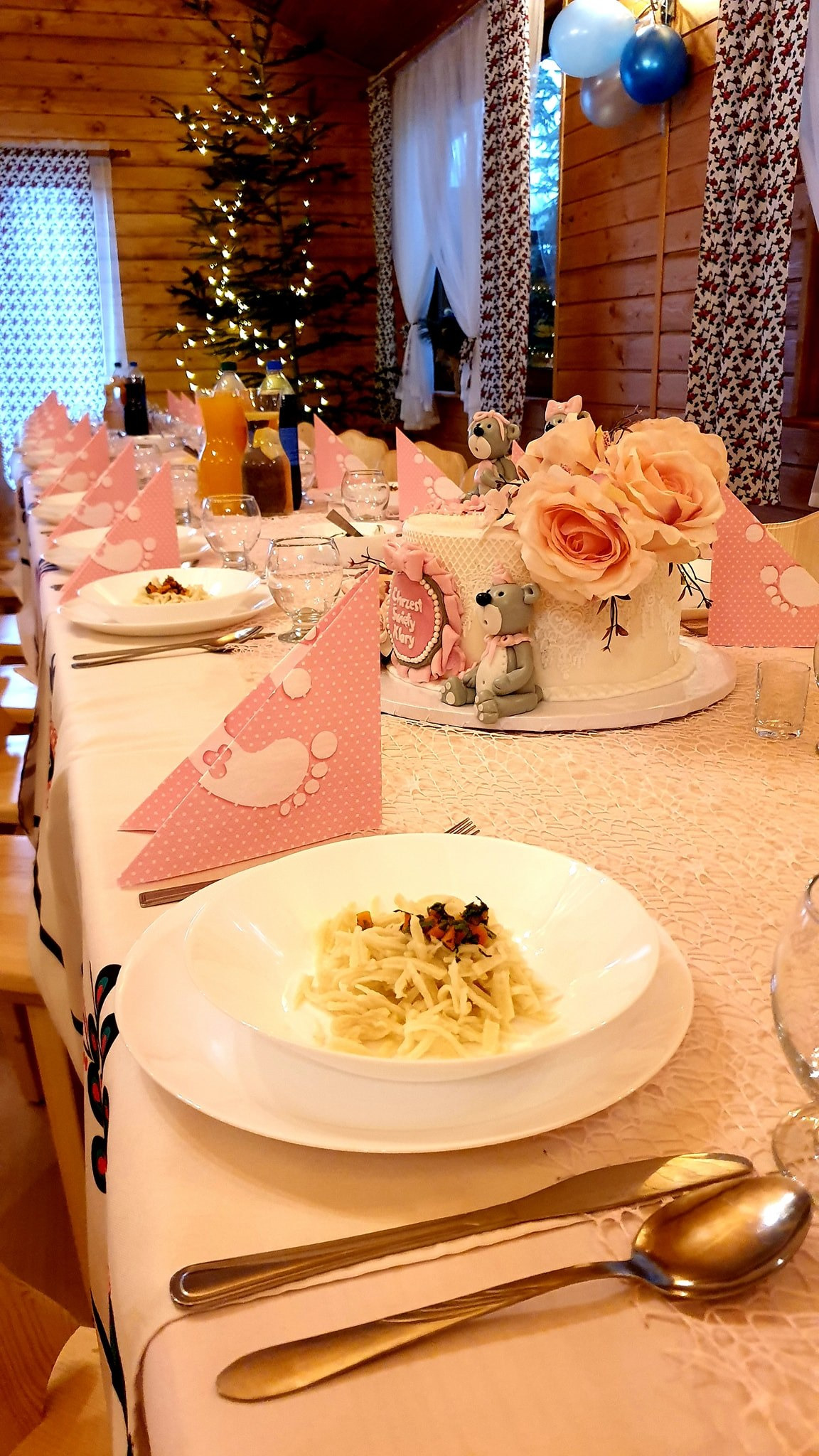 Uroczysty stół z białym obrusem, nakryty dla wielu osób, z dekoracjami w różowych odcieniach, tortem ozdobionym różami i figurkami misiów, przygotowany na przyjęcie z okazji Chrztu Świętego.