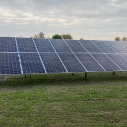 Dwa rzędy paneli słonecznych na metalowej konstrukcji, ustawione na trawiastym terenie z widokiem na dom i drzewa w tle pod pochmurnym niebem.