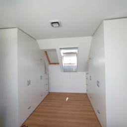 Też tak chcę - Wnętrze poddasza z białymi szafami w zabudowie, drewnianą podłogą i oknem dachowym z roletą.