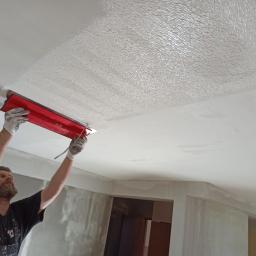 Też tak chcę - Pracownik w kombinezonie szpachluje sufit czerwoną szpachelką, pozostawiając widoczną fakturę. Częściowo wyszpachlowany sufit w trakcie prac wykończeniowych.