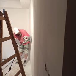 Też tak chcę - Mężczyzna w czerwonej koszulce i czapce z daszkiem szlifuje ścianę za pomocą szlifierki elektrycznej, stojąc na drabine w pomieszczeniu w trakcie remontu. Widoczne otwory po gniazdkach elektrycznych.