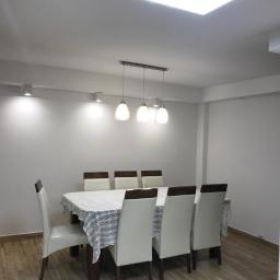 Też tak chcę - Jasne wnętrze jadalni w Rzeszowie po odświeżeniu ścian, z drewnianą podłogą i białymi krzesłami wokół stołu nakrytego koronkowym obrusem, oświetlone trzema wiszącymi lampami i punktowymi reflektorami.