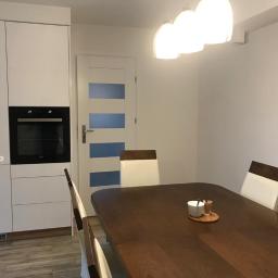 Też tak chcę - Nowoczesna jadalnia: białe szafki kuchenne z wbudowanym piekarnikiem, drewniany stół z białymi krzesłami, białe drzwi z niebieskimi akcentami, wiszące lampy kuliste.