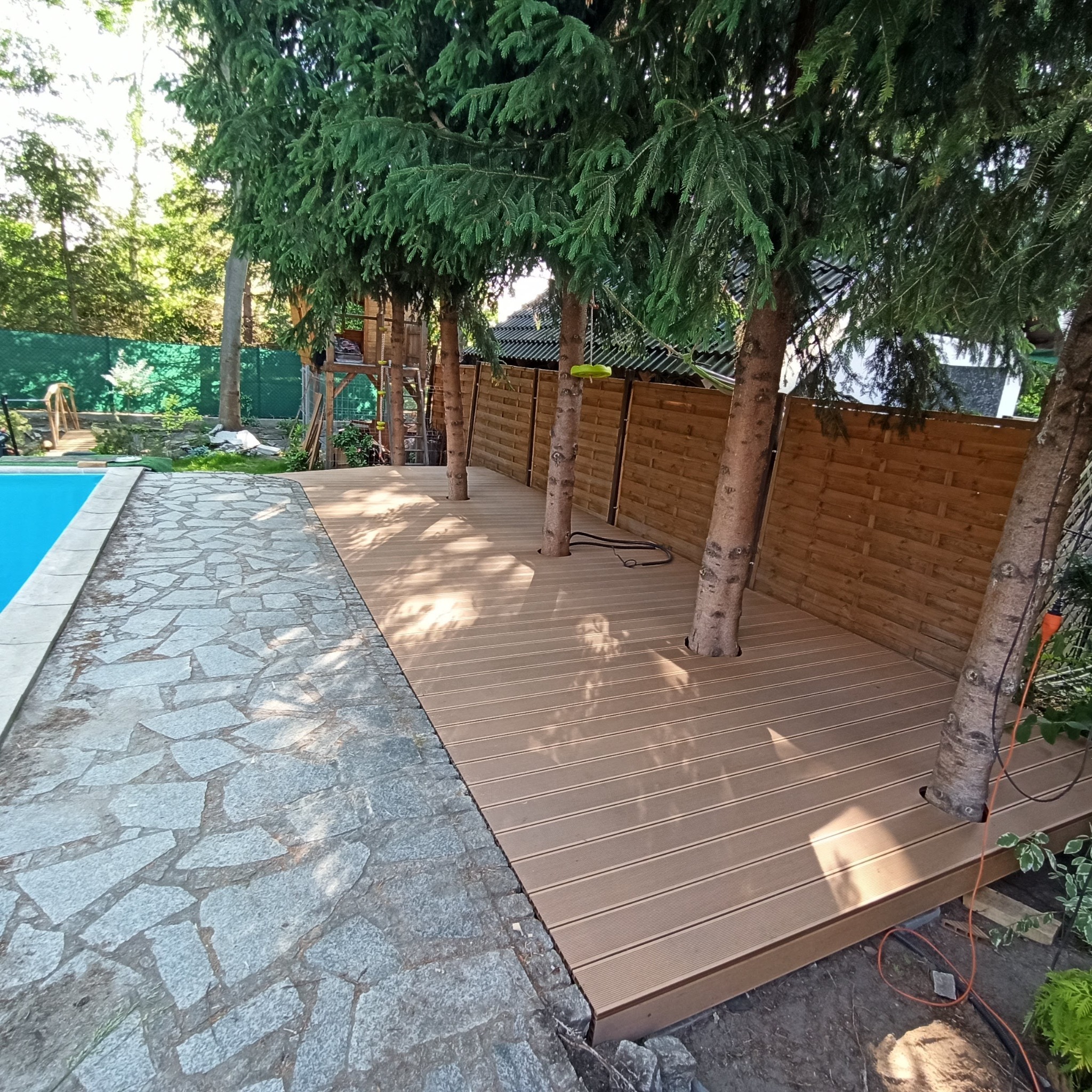 Kompozytowy taras z desek w kolorze jasnego drewna, zbudowany wokół rosnących drzew, obok basenu i kamiennej ścieżki.
