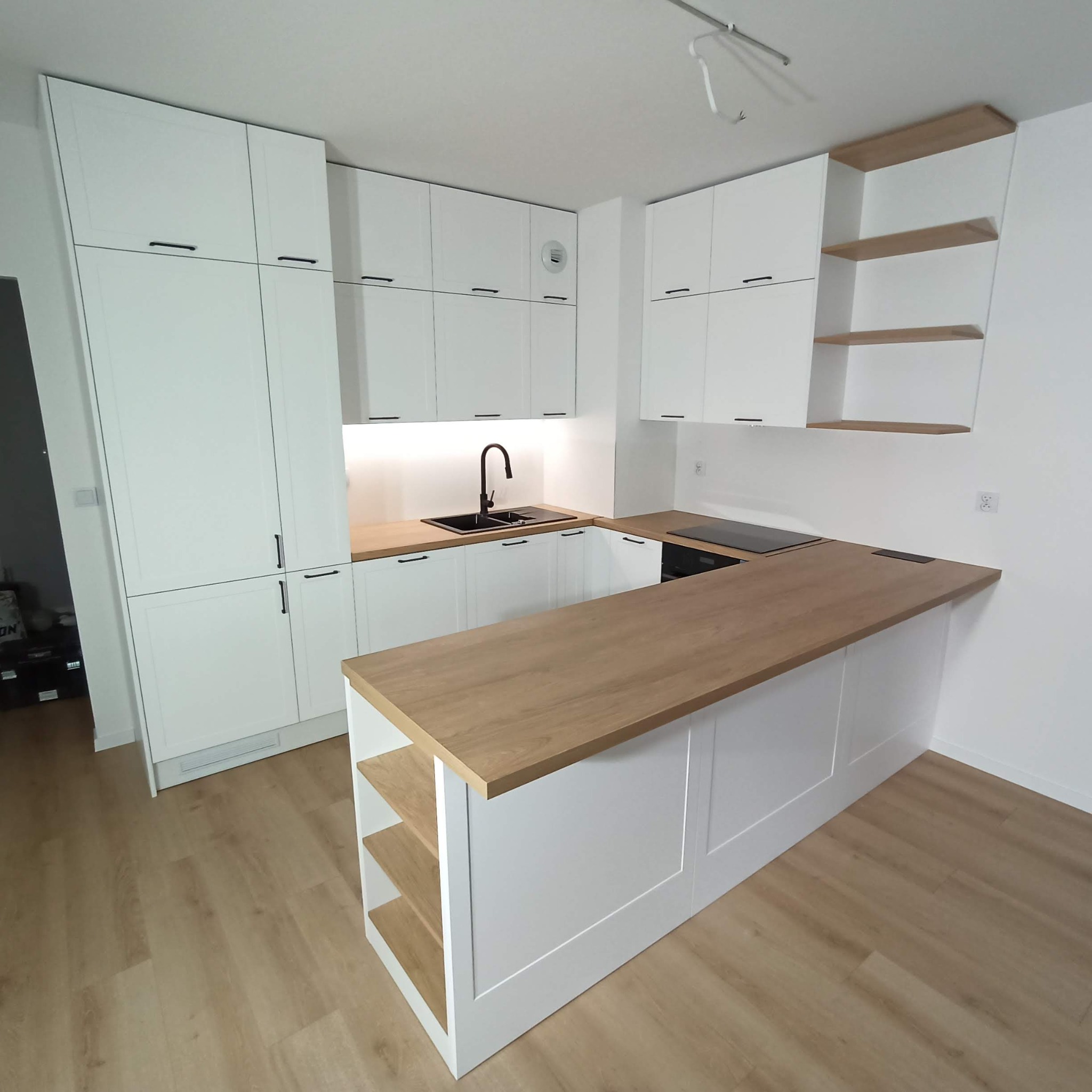 Nowoczesna, jasna kuchnia z białymi szafkami, drewnianym blatem i czarną armaturą, oświetlona listwą LED pod szafkami wiszącymi, z widocznymi panelami podłogowymi.
