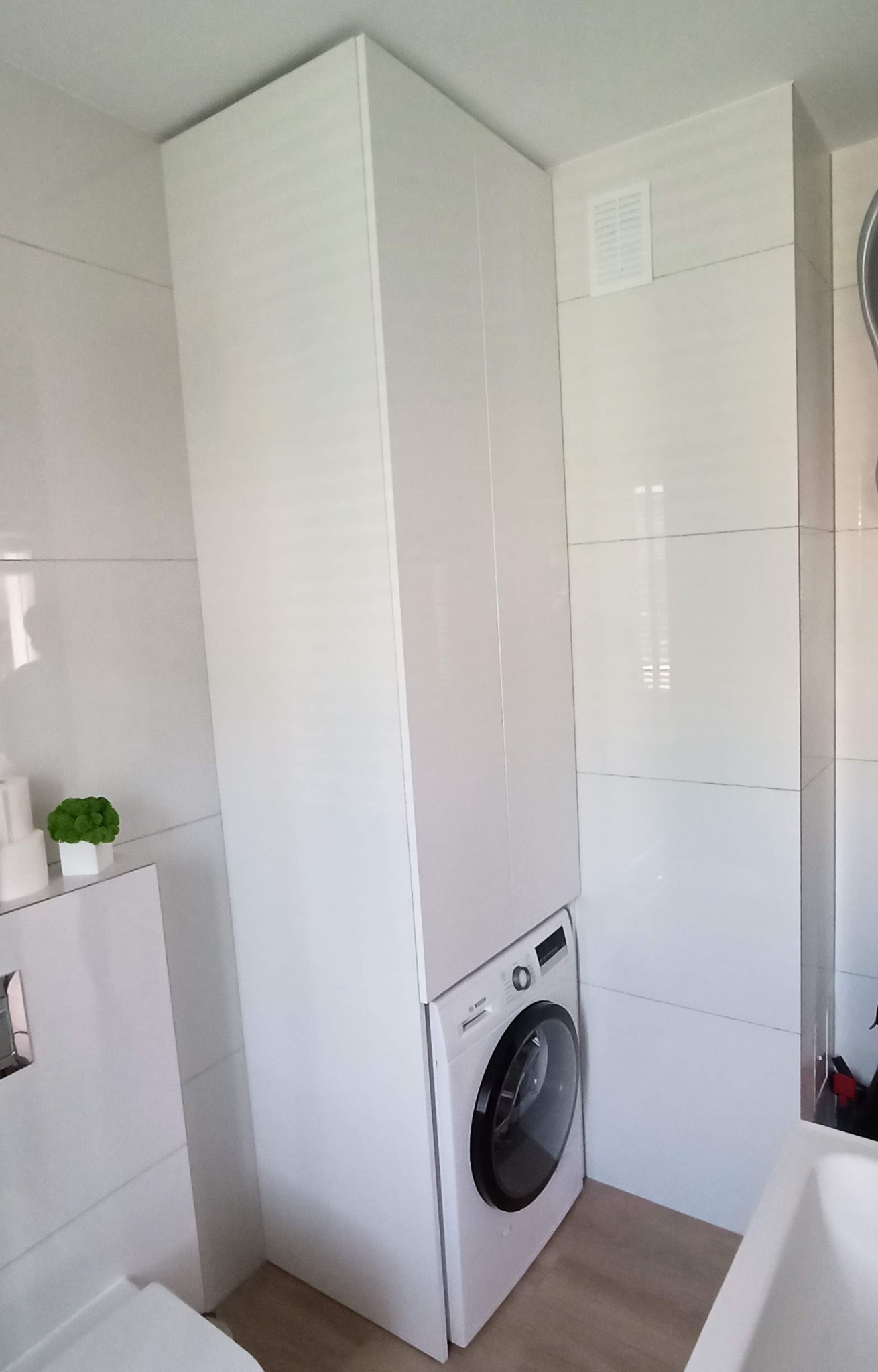 Biała łazienka z pralką zabudowaną w wysokiej szafce, widoczne płytki na ścianach i jasna podłoga.
