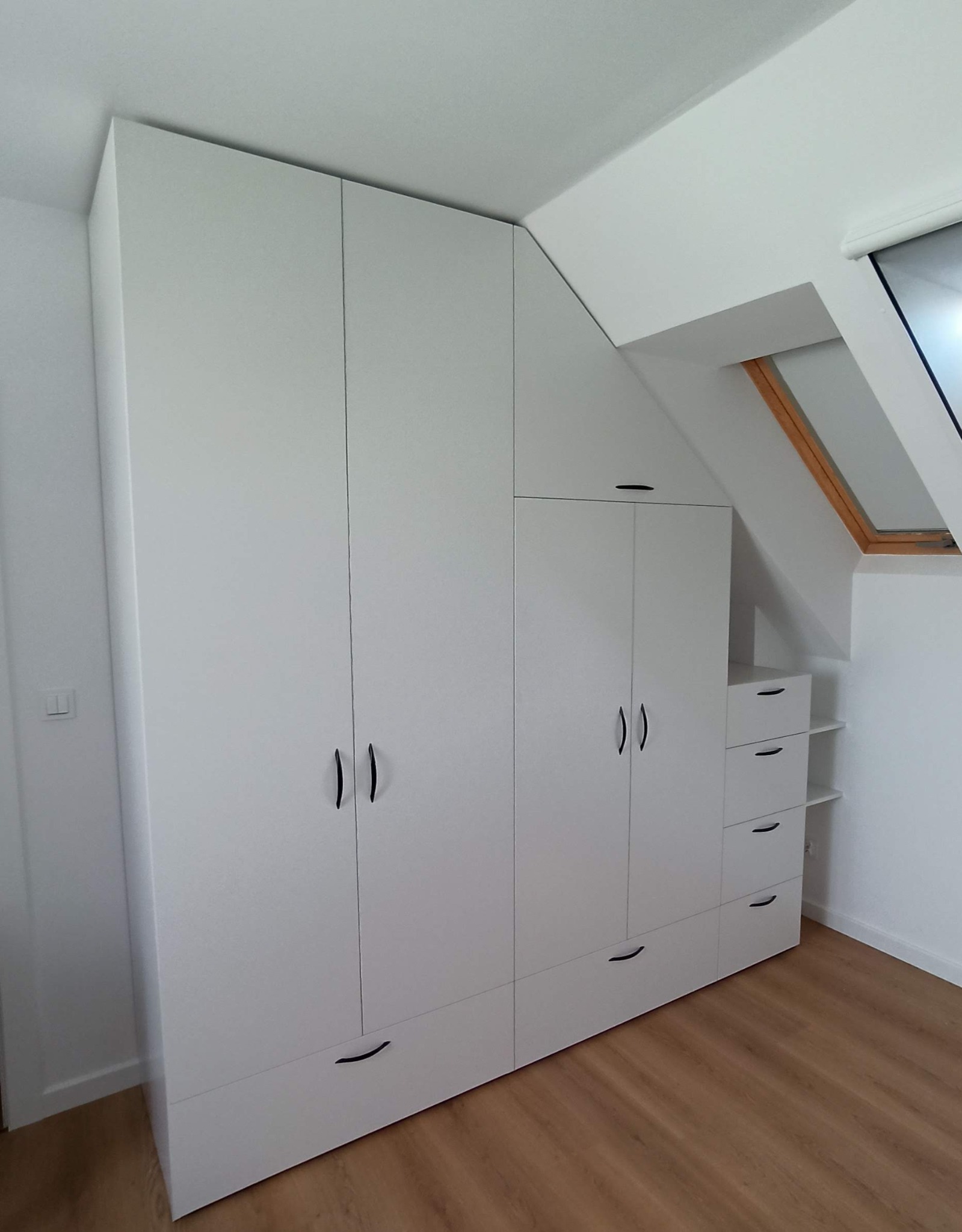 Biała zabudowa meblowa na wymiar, dopasowana do skosu dachu, z szafkami, szufladami i półkami, na podłodze panele podłogowe.