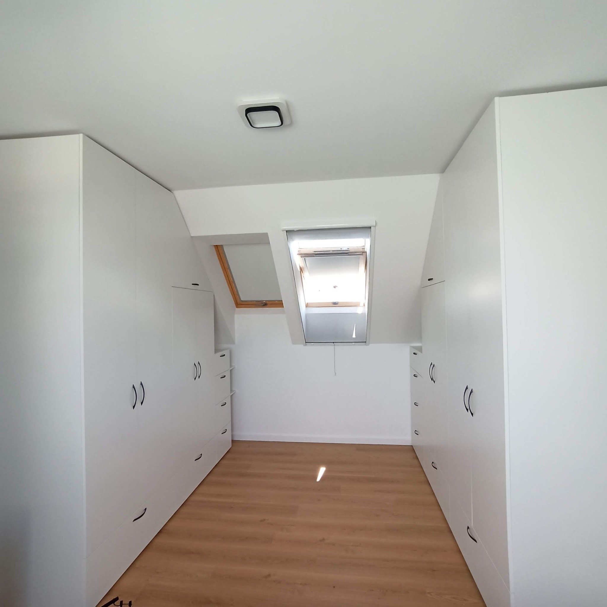 Wnętrze poddasza z białymi szafami w zabudowie, drewnianą podłogą i oknem dachowym z roletą.