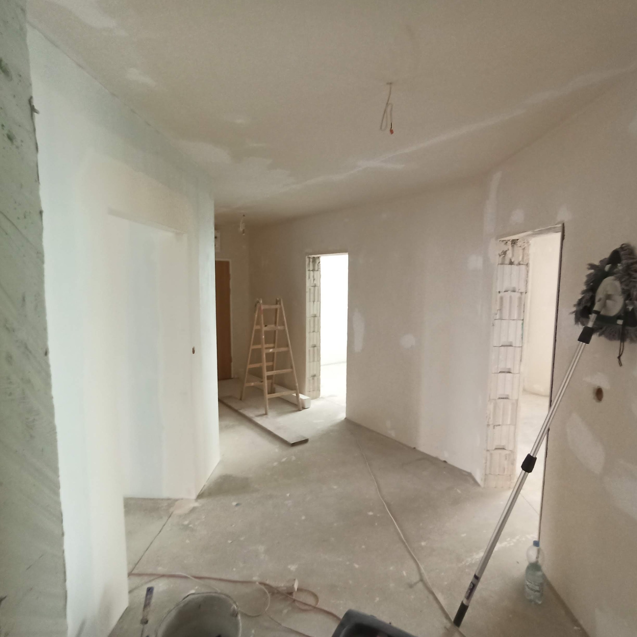 Wnętrze w trakcie szpachlowania ścian, widoczna drabina, narzędzia i butelka wody, surowe wykończenie ościeżnic.