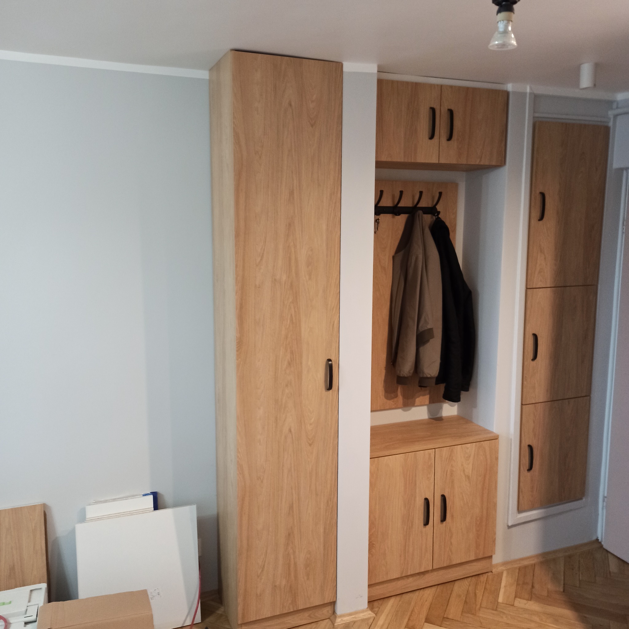 Zabudowa przedpokoju z szafami i wieszakiem na ubrania, wykończona okleiną drewnopodobną w odcieniu jasnego dębu, na podłodze parkiet w jodełkę.