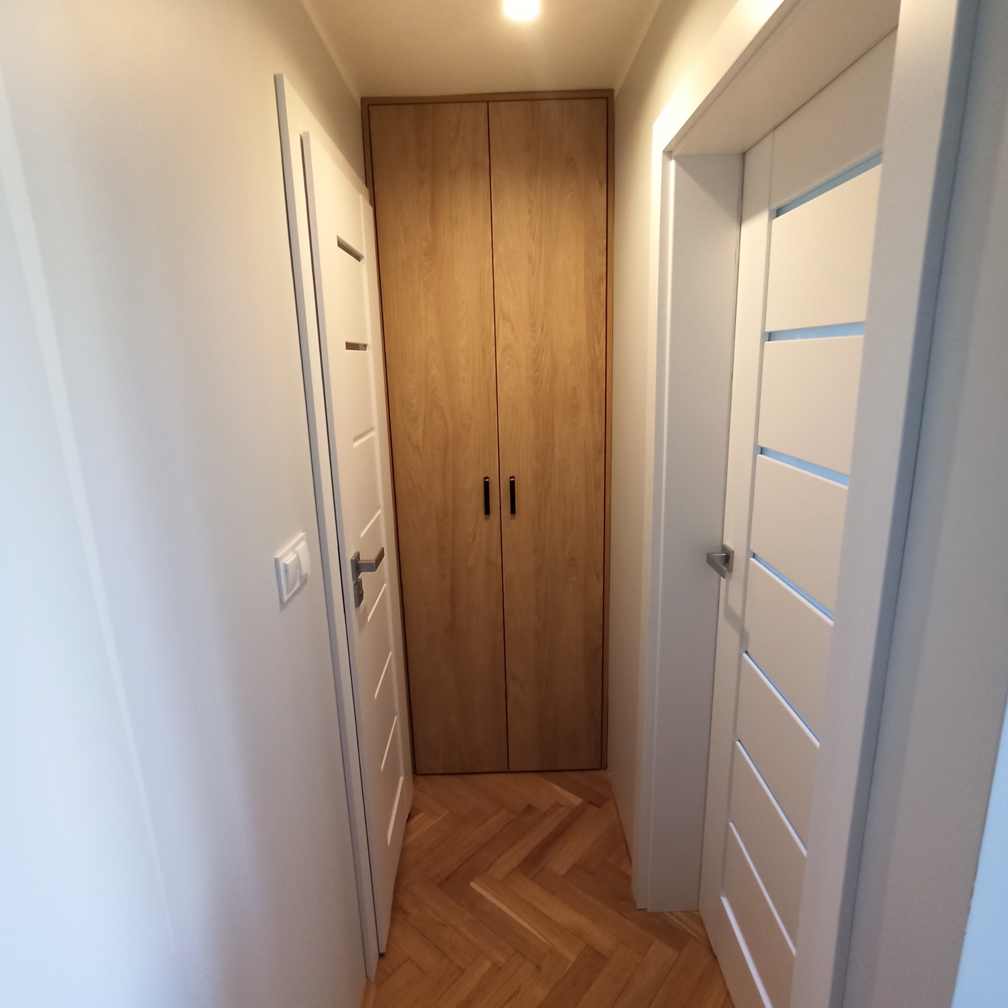 Korytarz z parkietem w jodełkę, białymi drzwiami z poziomymi frezami i drewnianą szafą w zabudowie z czarnymi uchwytami.