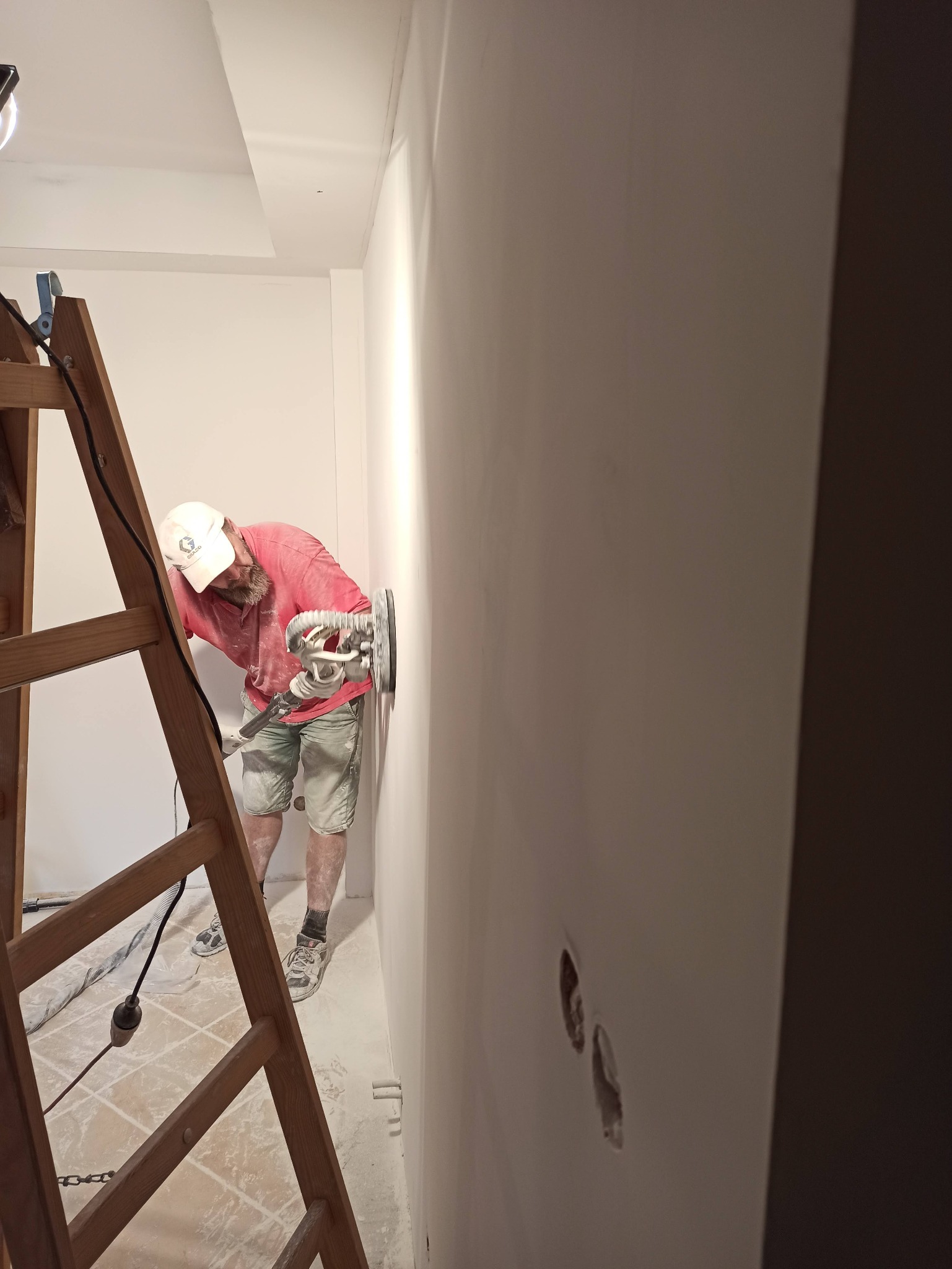 Mężczyzna w czerwonej koszulce i czapce z daszkiem szlifuje ścianę za pomocą szlifierki elektrycznej, stojąc na drabine w pomieszczeniu w trakcie remontu. Widoczne otwory po gniazdkach elektrycznych.