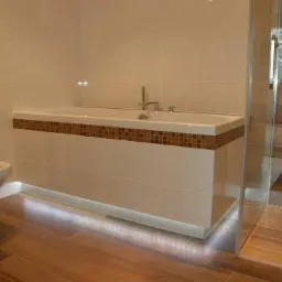 Nowoczesna łazienka z podświetlaną wanną obudowaną płytkami, mozaika w odcieniach brązu na obudowie wanny i ścianie, kabina prysznicowa ze szklanymi drzwiami, toaleta z otwartą klapą, podłoga...