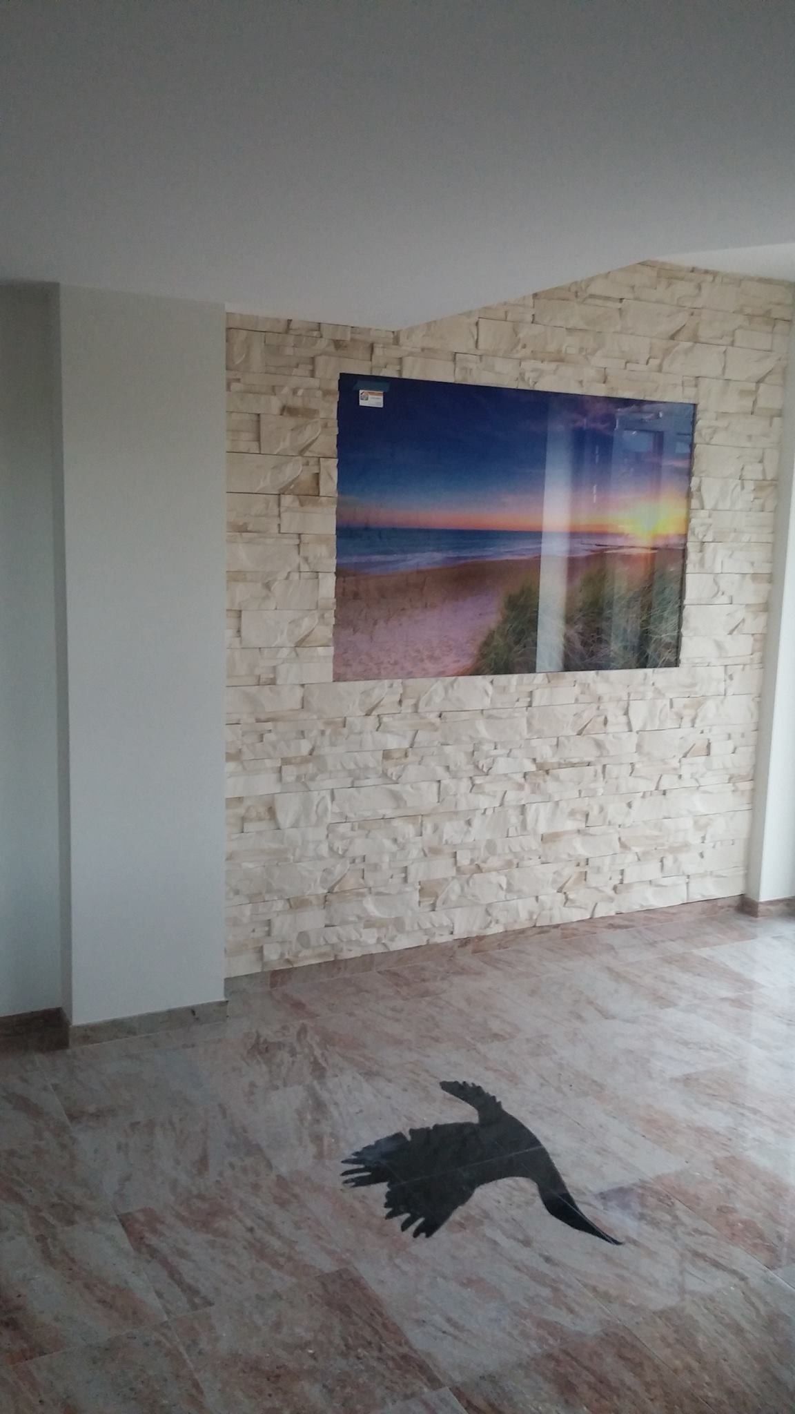 Wykończone wnętrze: ściana z dekoracyjnej, jasnej cegły z naklejonym dużym plakatem przedstawiającym zachód słońca na plaży, marmurowa podłoga z czarną sylwetką ptaka.