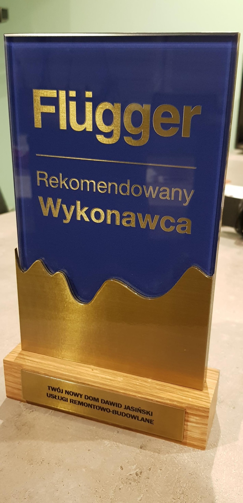 Szklana statuetka 'Rekomendowany Wykonawca' marki Flügger na drewnianej podstawie z tabliczką 'Twój Nowy Dom Dawid Jasiński Usługi Remontowo-Budowlane'.