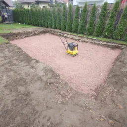 Przygotowany wykop pod budowę z zagęszczoną warstwą podkładową; widoczna zagęszczarka wibrująca na środku i rząd tuj w tle.