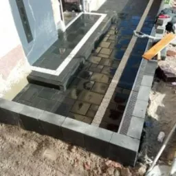 Nowo ułożona, ciemna kostka brukowa przed budynkiem, z jasnymi paskami oddzielającymi fragmenty, oraz z zamontowanym odpływem liniowym.