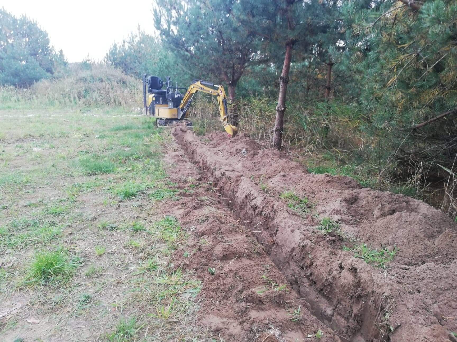 Żółta mini koparka na skraju lasu, tworząca wąski wykop w ziemi, widoczne ślady opon i świeżo wykopana ziemia.