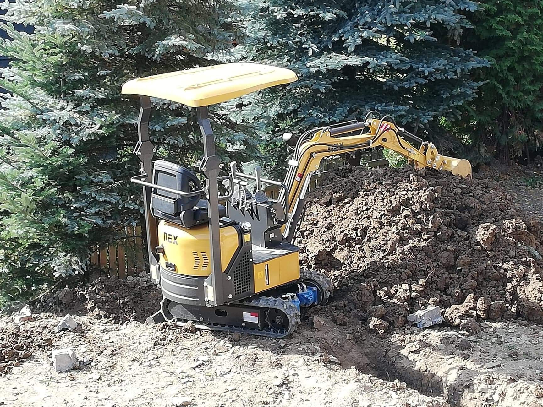 Żółta mini koparka NEX w trakcie pracy, usypuje brązową ziemię obok wykopanego wąskiego rowu na tle zielonych drzew iglastych.