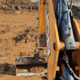 Żółta koparka JCB w trakcie prac ziemnych, widoczny wykop w grząskiej, błotnistej ziemi, w tle budynek mieszkalny.