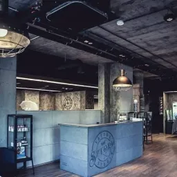 Wnętrze nowoczesnego salonu fryzjerskiego z betonową recepcją z logo, oświetleniem w stylu industrialnym i stanowiskami pracy w tle.