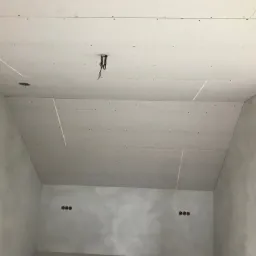 Poddasze z zamontowanymi płytami kartonowo-gipsowymi, widoczne łączenia, otwór na oświetlenie i wystające przewody, ściany w stanie surowym przed malowaniem.