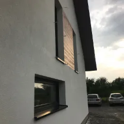 Boczny widok na otynkowaną ścianę budynku z dwoma oknami; jedno okno osłonięte płytami, widoczne dwa samochody w tle i pochmurne niebo.