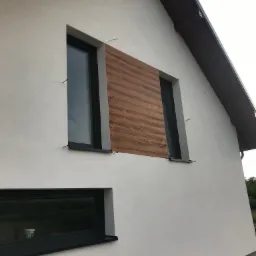 Elewacja domu jednorodzinnego w Łańcucie z widocznym ociepleniem fragmentu ściany zewnętrznej imitacją drewna, obok okien z czarnymi ramami. Widoczne wystające druty montażowe.
