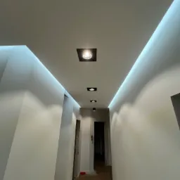 Nowoczesny korytarz z białymi ścianami, drewnianą podłogą i podwieszanym sufitem z oświetleniem LED oraz punktowymi lampami, widok w perspektywie.