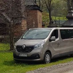 Szary Renault Trafic zaparkowany na trawie z ż&oacute;łtymi kwiatami, na tle drewnianej architektury sakralnej.