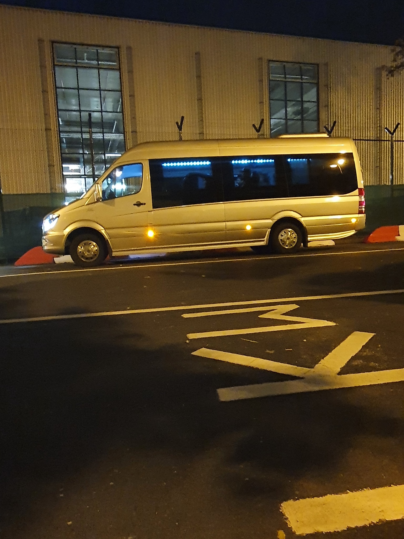 Złoty bus zaparkowany nocą przy budynku z dużymi oknami, widoczne oznaczenia poziome na asfalcie.