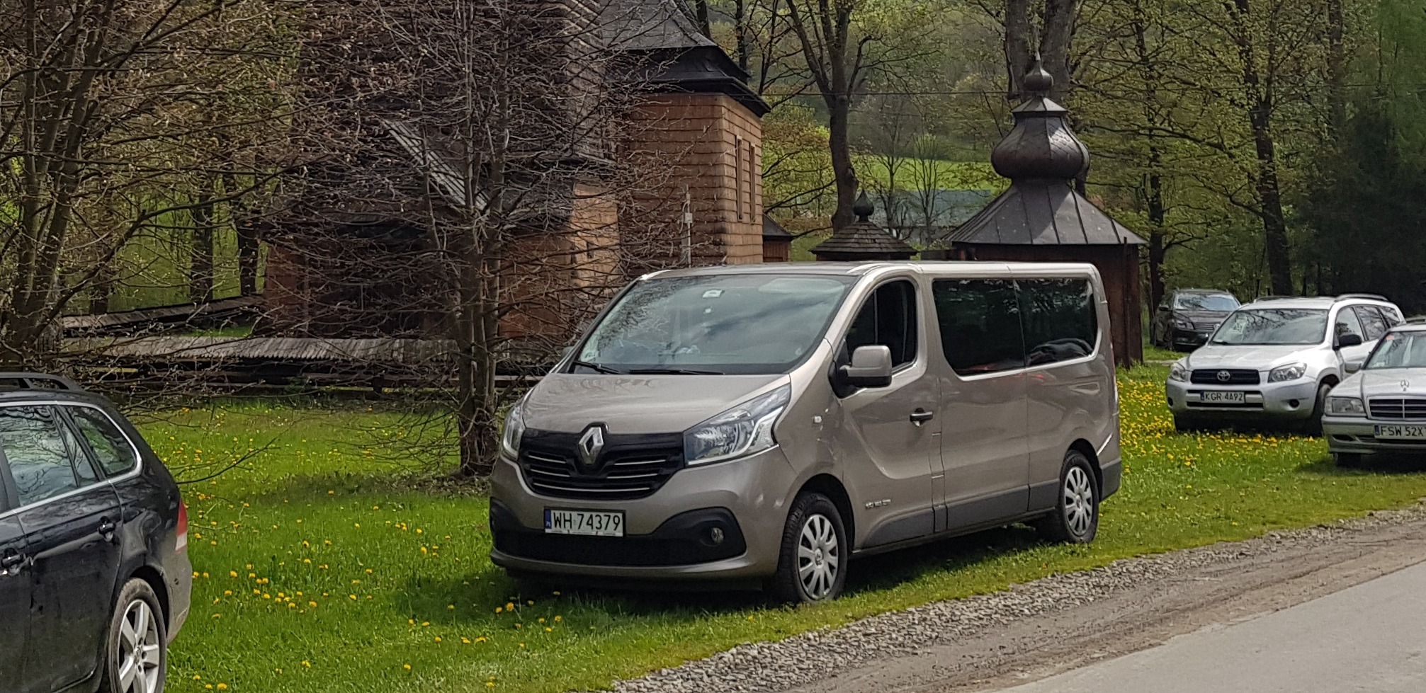 Szary Renault Trafic zaparkowany na trawie z żółtymi kwiatami, na tle drewnianej architektury sakralnej.