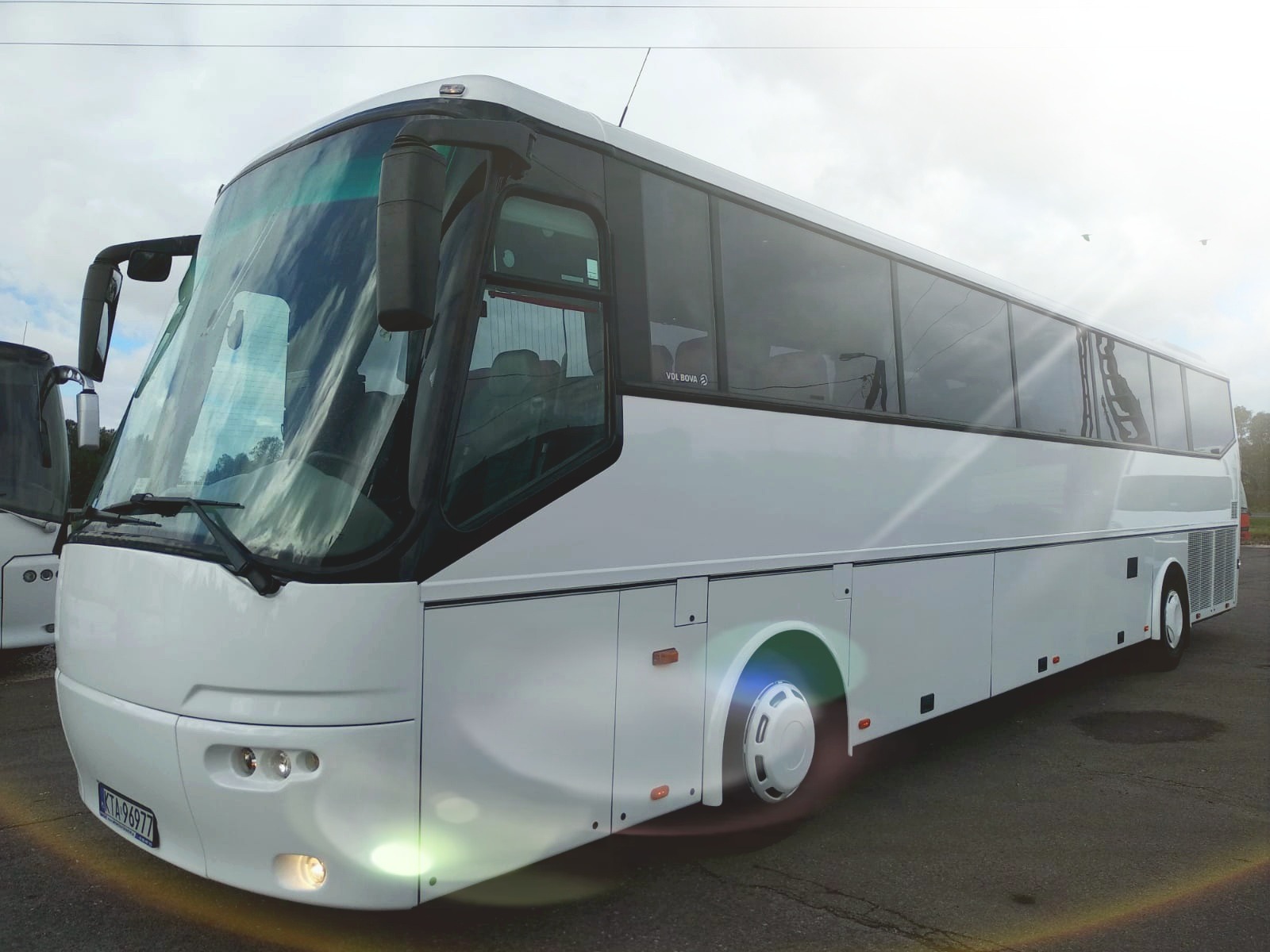 Biały autokar turystyczny marki VDL BOVA, zaparkowany na zewnątrz, z widocznymi odbiciami w szybach i włączonymi światłami mijania.
