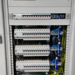 Otwarta, biała szafa elektryczna z zainstalowanymi bezpiecznikami, przewodami w kolorach niebieskim, żółto-zielonym i brązowym, widoczne elementy montażowe i szyny DIN.