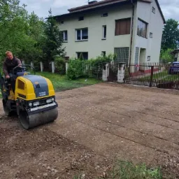 Operator na walcu drogowym zagęszcza grunt na podwórku przed domem jednorodzinnym z metalową bramą w tle.
