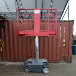 Podnośnik koszowy pionowy, czerwona platforma robocza, szara podstawa na kołach, na zewnątrz na kostce brukowej, w tle brązowy kontener i panele słoneczne na dachu.