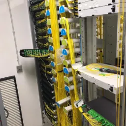 Panel krosowniczy z uporządkowanymi kablami światłowodowymi oznaczonymi etykietami A-L w serwerowni, widoczne narzędzia do światłowodów na półce.