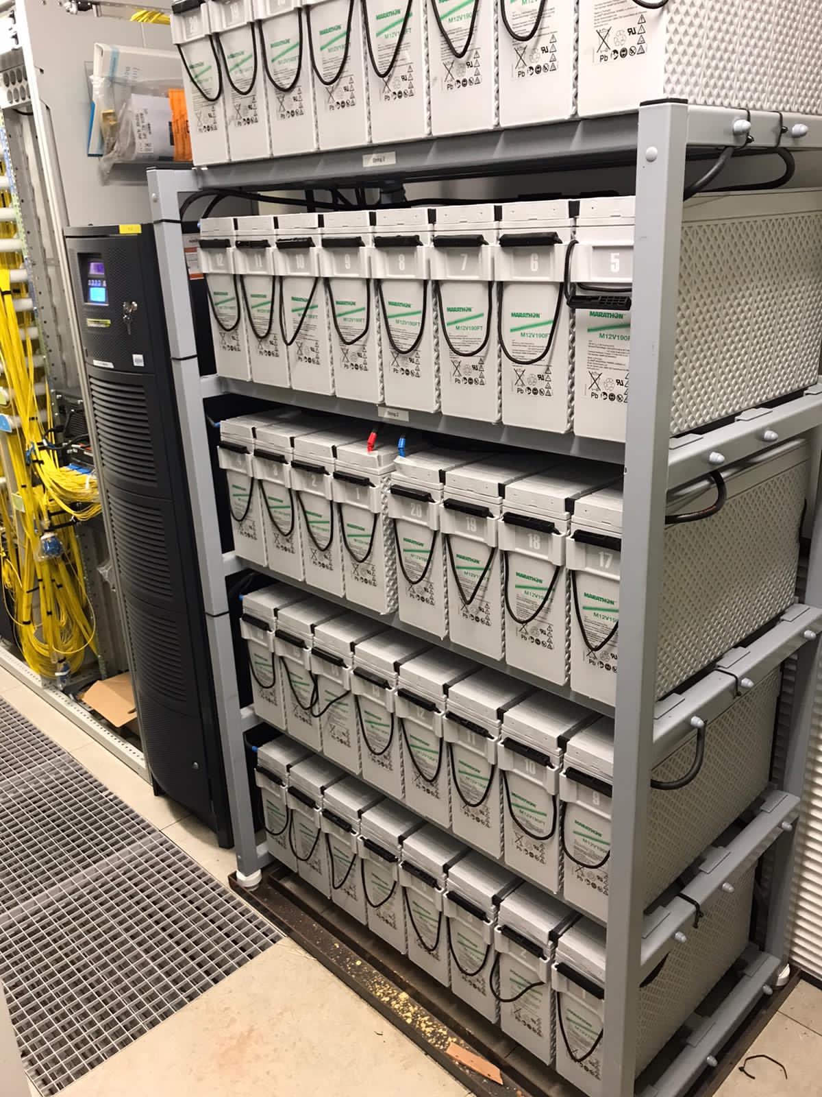 Bateria akumulatorów Marathon M12V190FT ułożona na metalowym regale w serwerowni obok szafy rack z okablowaniem.