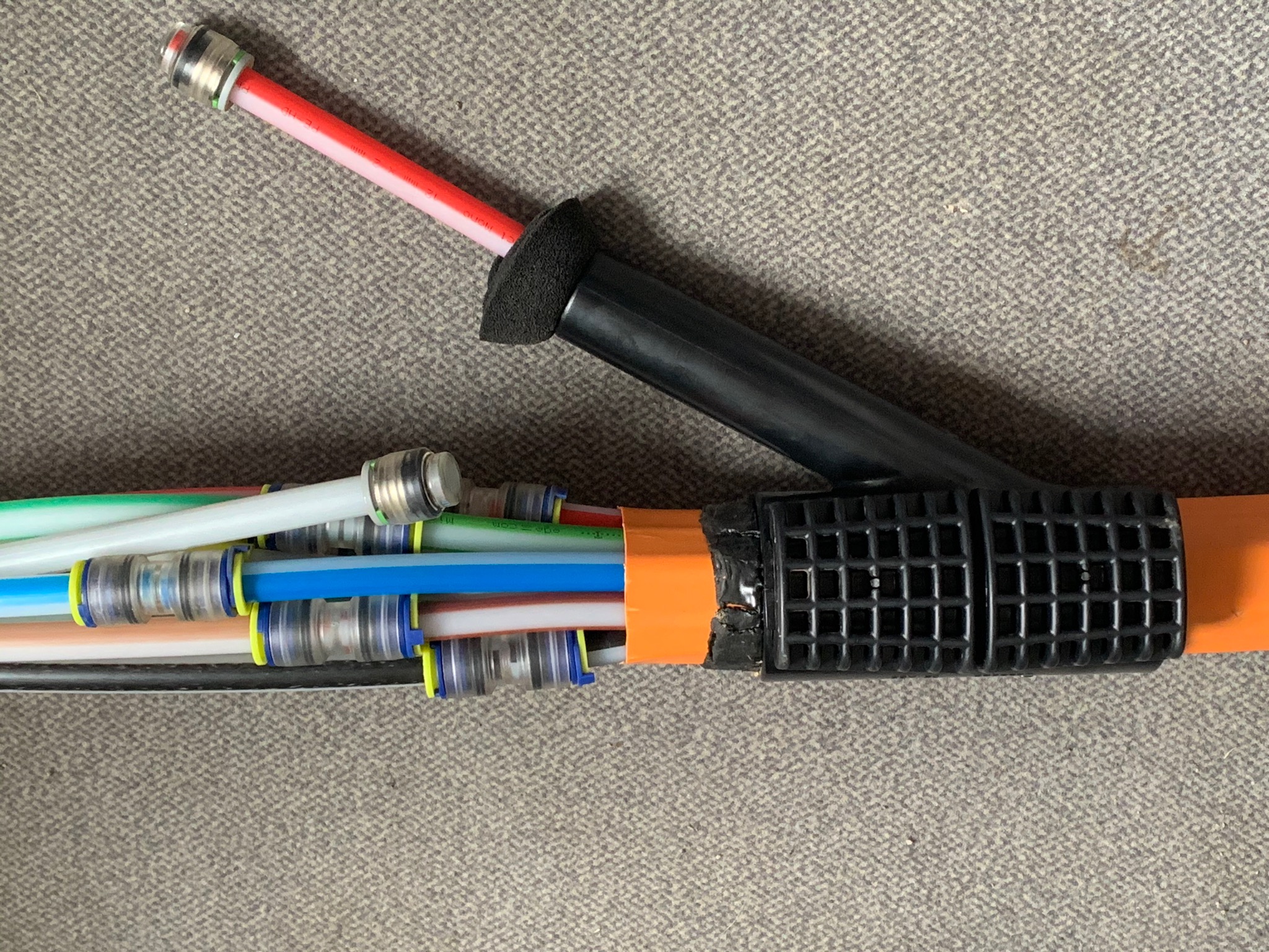 Zestaw kolorowych rurek instalacyjnych z metalowymi złączkami, połączonych w wiązkę i zabezpieczonych pomarańczową taśmą oraz czarną, ażurową osłoną, leżący na szarym dywanie.