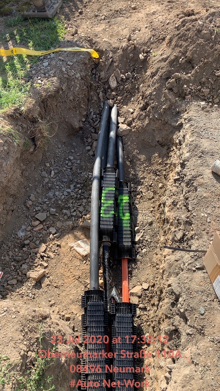 Wykop z trzema czarnymi rurami kablowymi zabezpieczonymi czarnymi osłonami z zielonymi oznaczeniami, ułożonymi w ziemi z żółtą taśmą ostrzegawczą w tle.