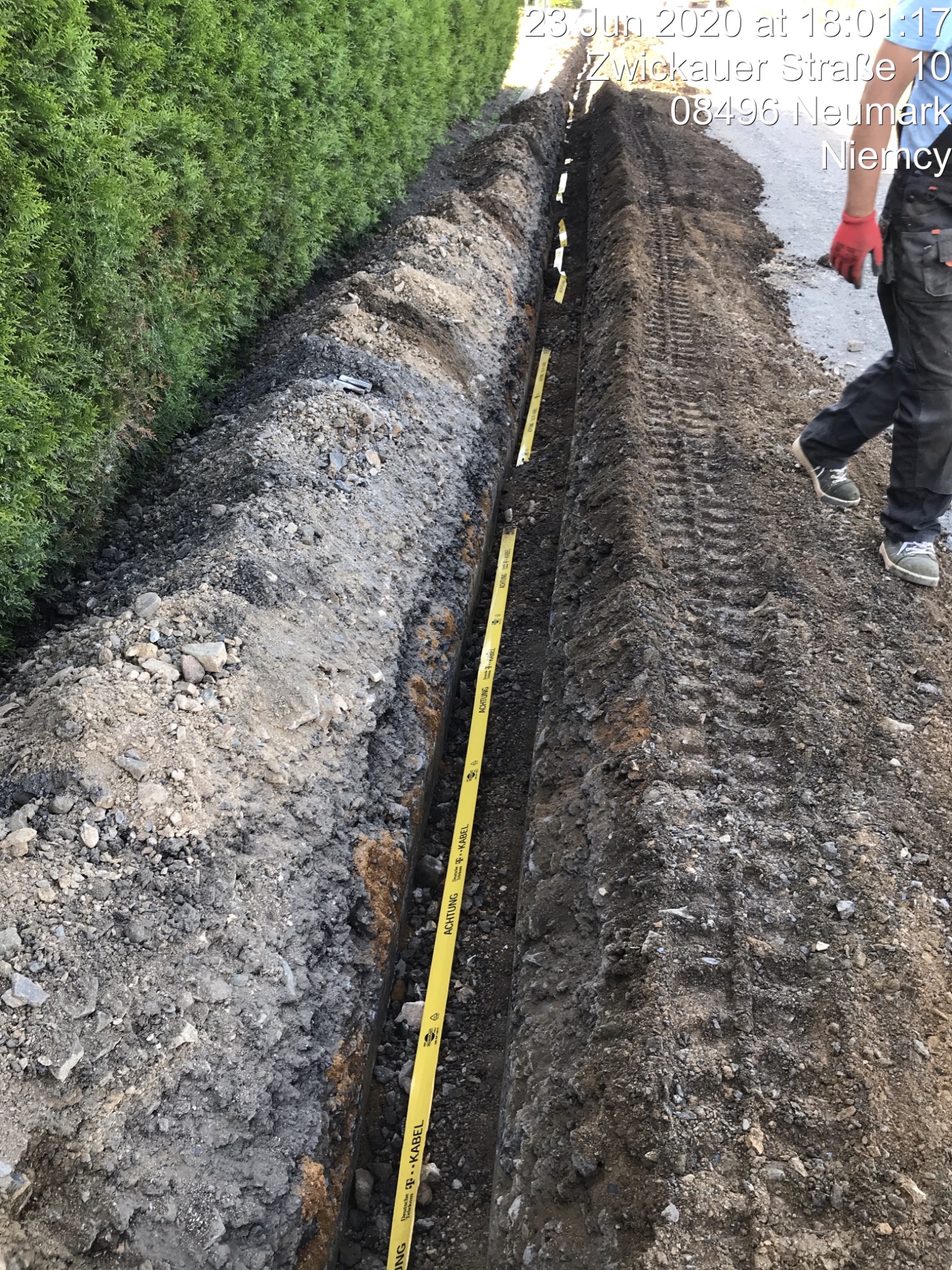 Wykop w ziemi z żółtą taśmą ostrzegawczą Deutsche Telekom Kabel, widoczne ślady opon i fragment sylwetki osoby w czerwonych rękawicach.