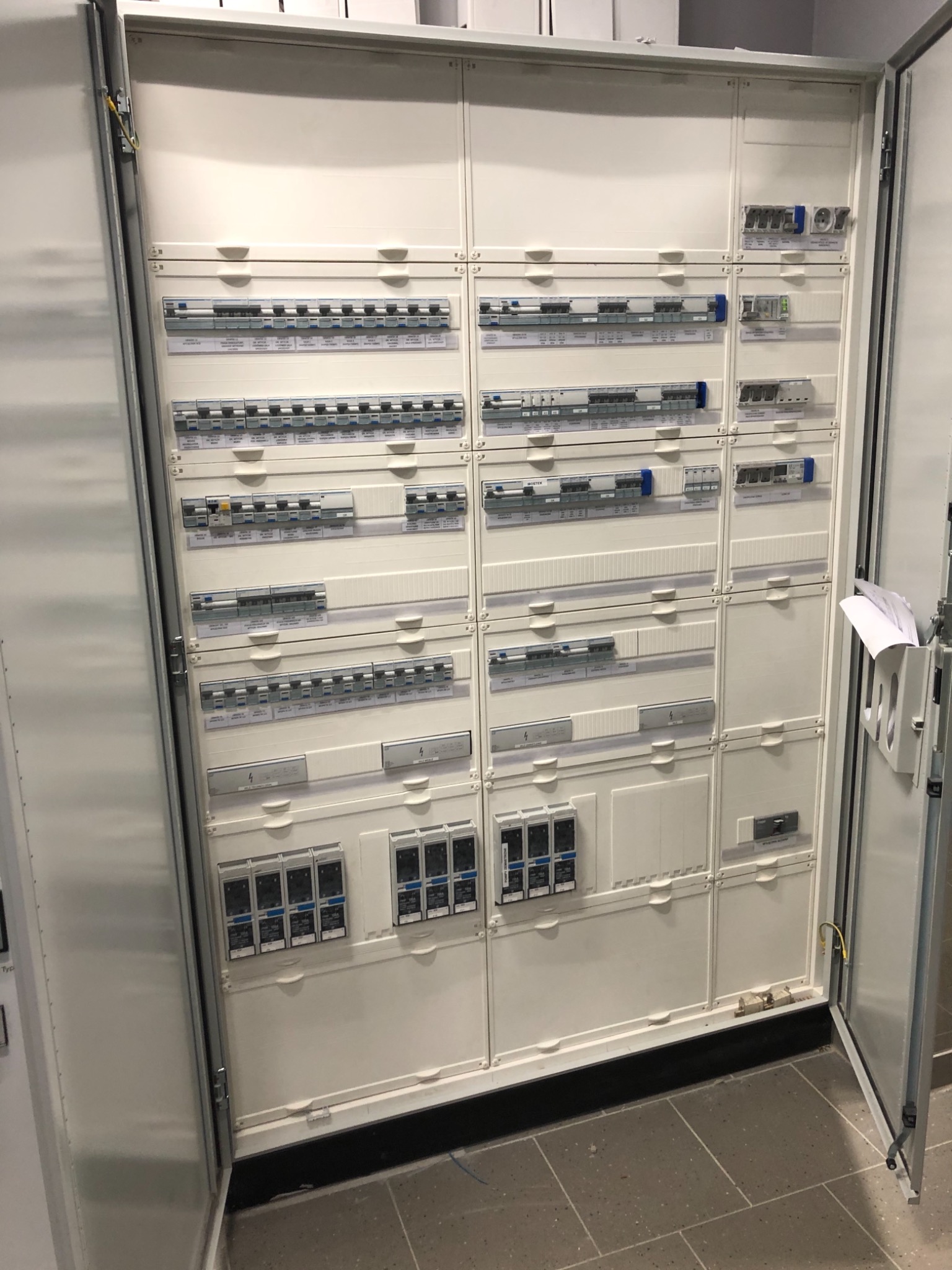Otwarta, biała szafa rozdzielcza z zamontowanymi bezpiecznikami i wyłącznikami, widoczne etykiety opisowe, dokumentacja w uchwycie na drzwiczkach.