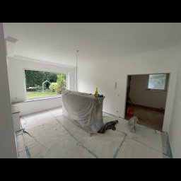Marcin Gąsiorowski Usługi Remontowo Budowlane - Pokój w trakcie remontu: świeżo pomalowane ściany na biało, podłoga zabezpieczona folią malarską, mebel zakryty folią ochronną, widoczne narzędzia i butelka na blacie.