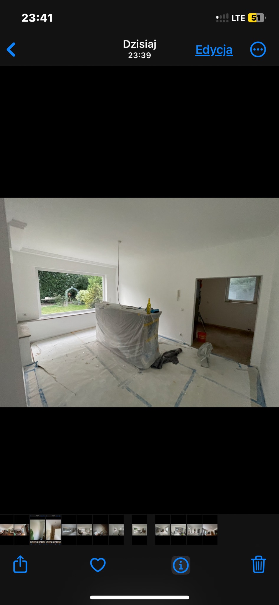 Pokój w trakcie remontu: świeżo pomalowane ściany na biało, podłoga zabezpieczona folią malarską, mebel zakryty folią ochronną, widoczne narzędzia i butelka na blacie.