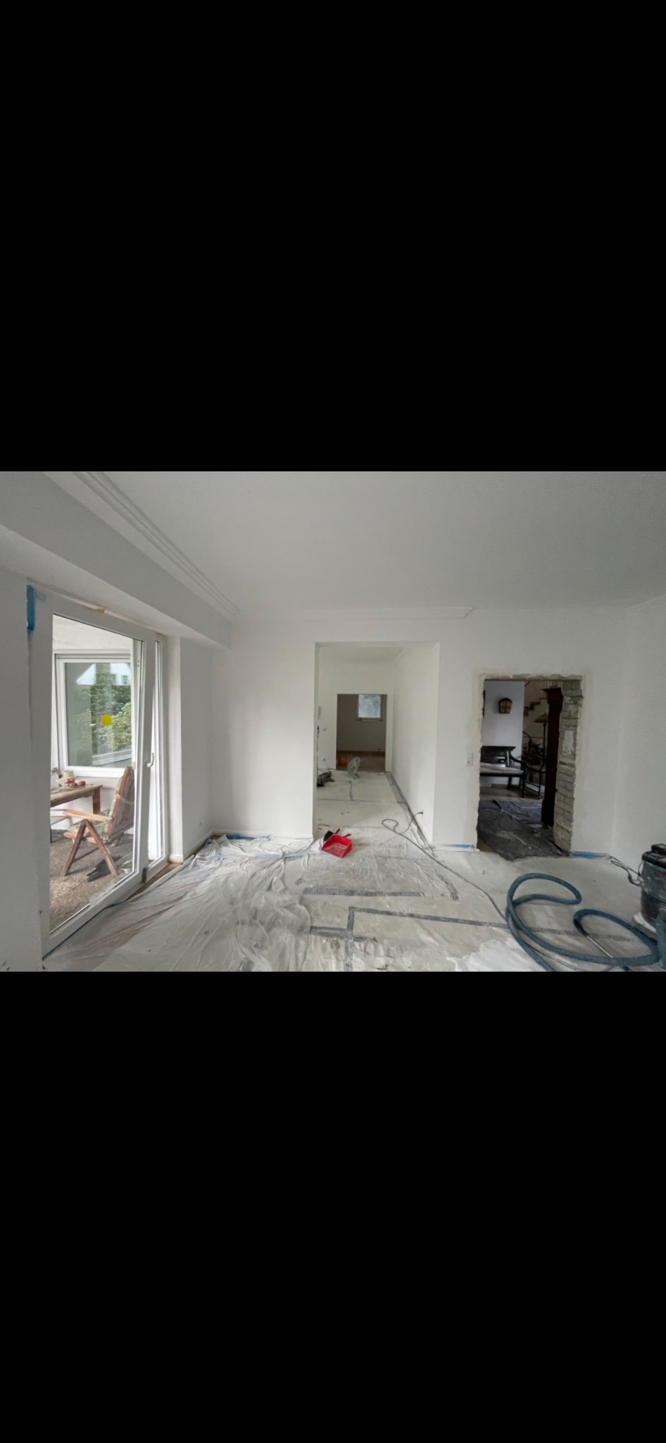 Pokój w trakcie remontu: ściany świeżo pomalowane na biało, podłoga zabezpieczona folią malarską, widoczne narzędzia i pozostałości po pracach murarskich przy jednym z przejść. Na zewnątrz widoczny...