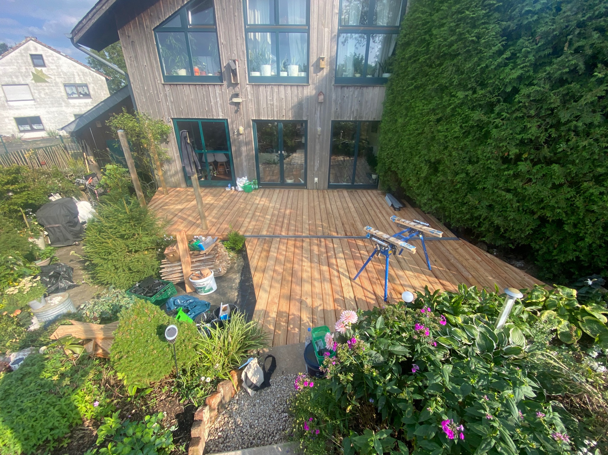 Nowa drewniana deska tarasowa przed domem, widok z góry, z widocznymi narzędziami, kwiatami i zielenią wokół.