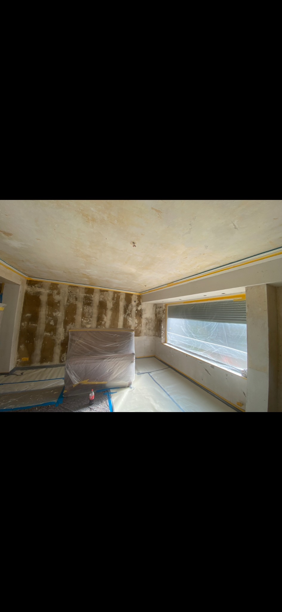 Pokój w trakcie przygotowania do remontu: ściany częściowo odarte z tapet, zabezpieczone folią okno, podłoga i meble przykryte folią ochronną, widoczne taśmy malarskie.