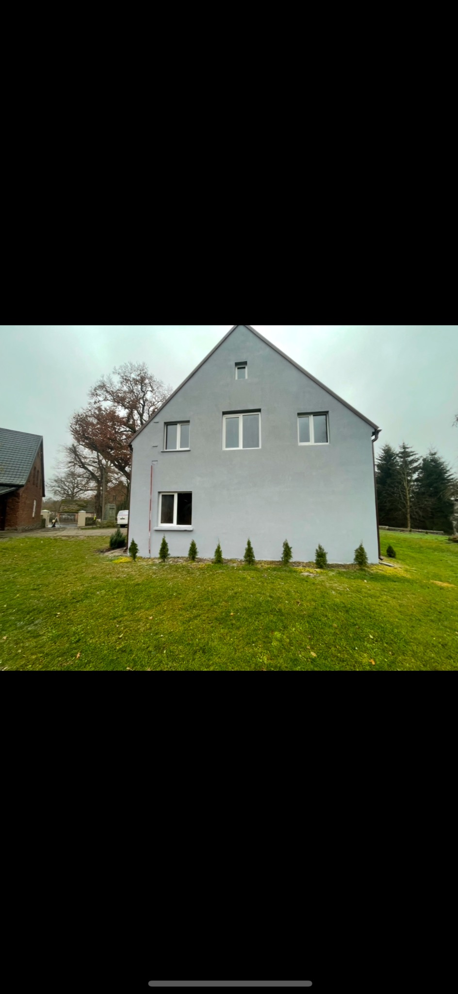 Odnowiony dom jednorodzinny z szarą elewacją, białymi ramami okiennymi i rzędem małych tujek posadzonych wzdłuż ściany, na tle zielonego trawnika i pochmurnego nieba.